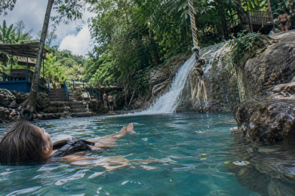 Swimmer floating in a bright blue natural pool beneath a small jungle waterfall