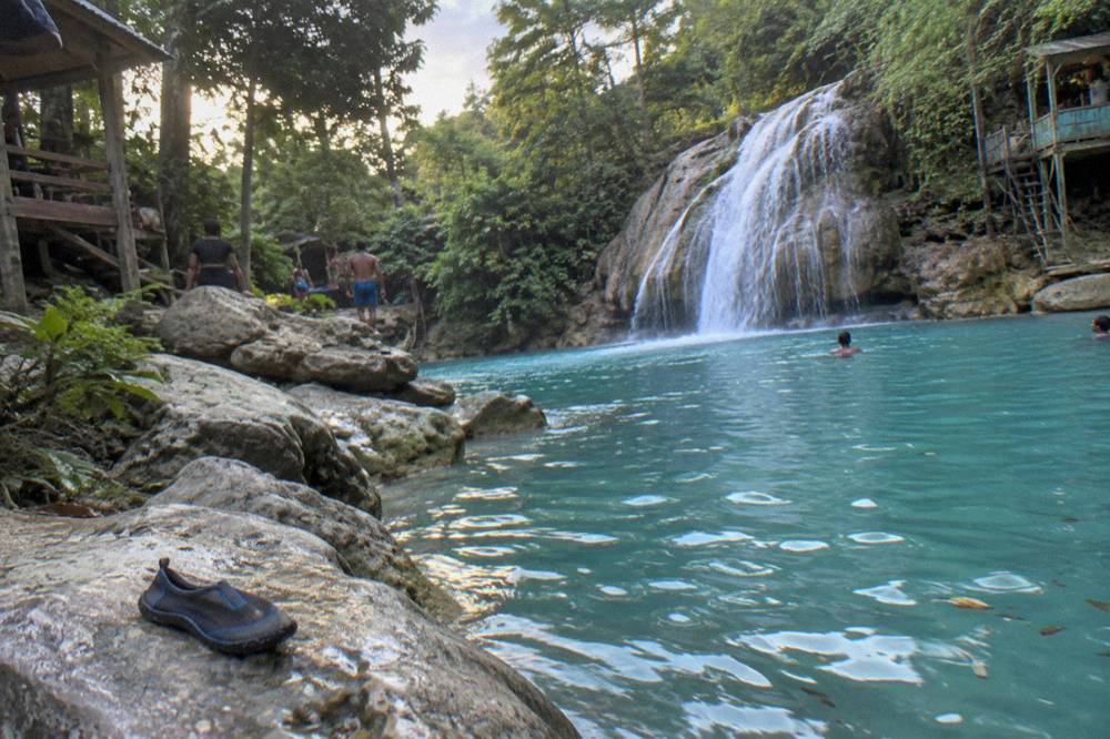 A calm turquoise pool at the base of a jungle waterfall with limestone rocks along the edge.