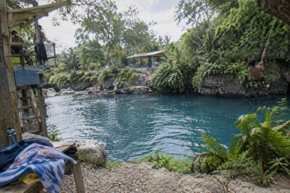 Swimmer on a rope swing above a blue limestone pool ringed by ferns