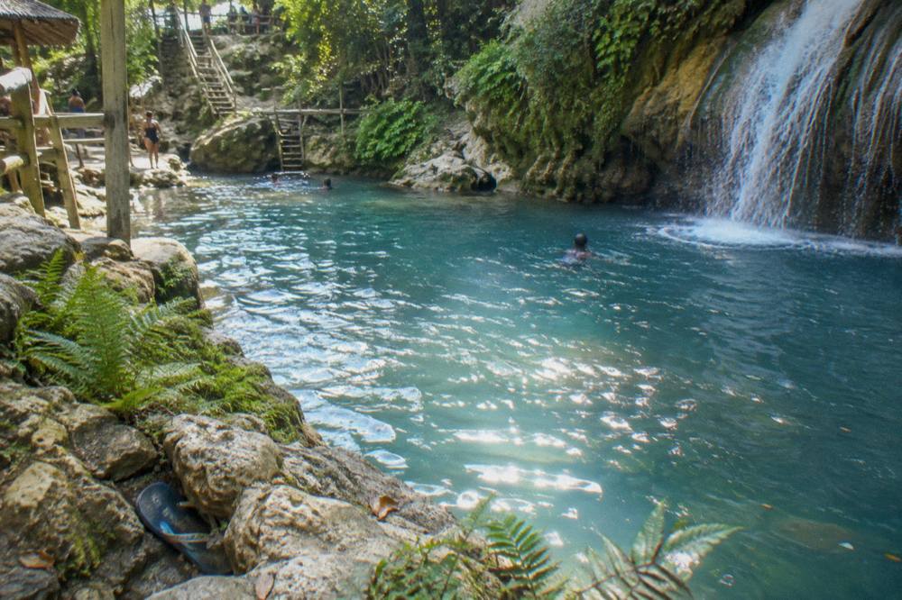 Sunlit waterfall pouring into a calm blue pool ringed by rocks and tropical trees