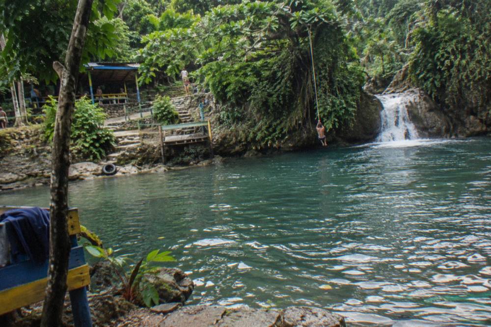Swimmer on a rope swing over a deep jungle pool below a small cascade
