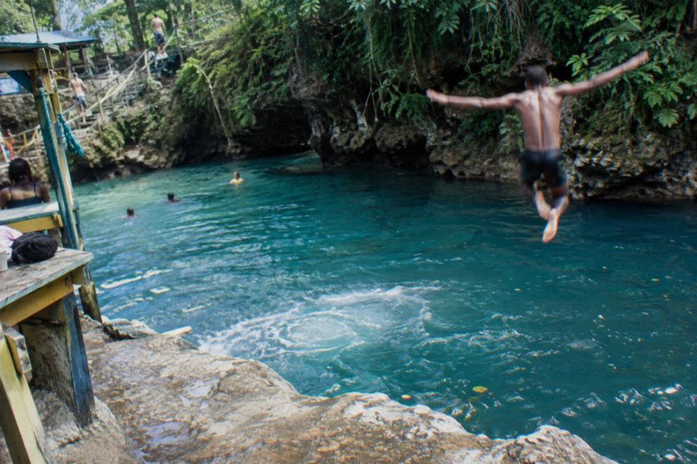 Swimmer mid-air leaping from a limestone ledge into a deep turquoise pool