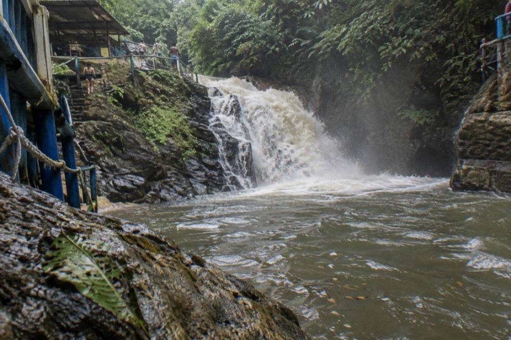 Waterfall rushing over dark rocks into a full green-tinted pool after rain