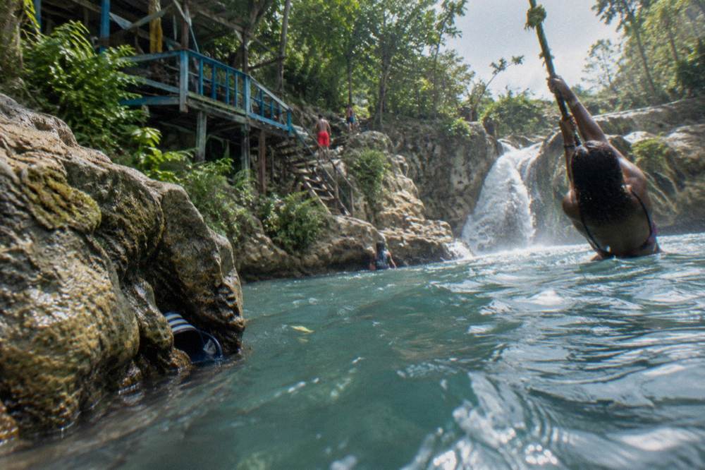 Swimmer holding a rope swing above a clear blue pool at a jungle waterfall