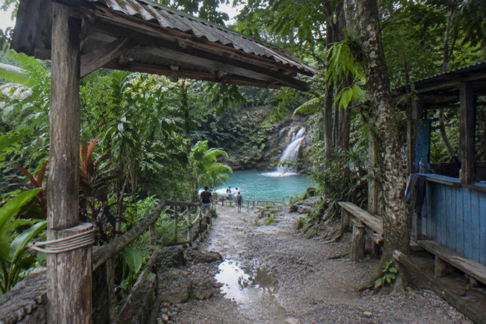 Wooden sign and jungle trail leading toward a turquoise waterfall pool