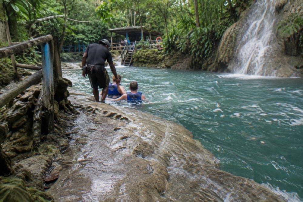Guide and visitors on a wet limestone path beside a waterfall pool