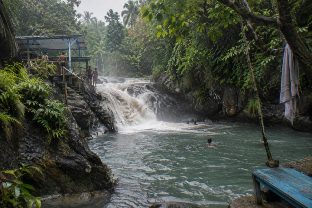 Waterfall rushing over dark rocks into a green-tinted pool after rain