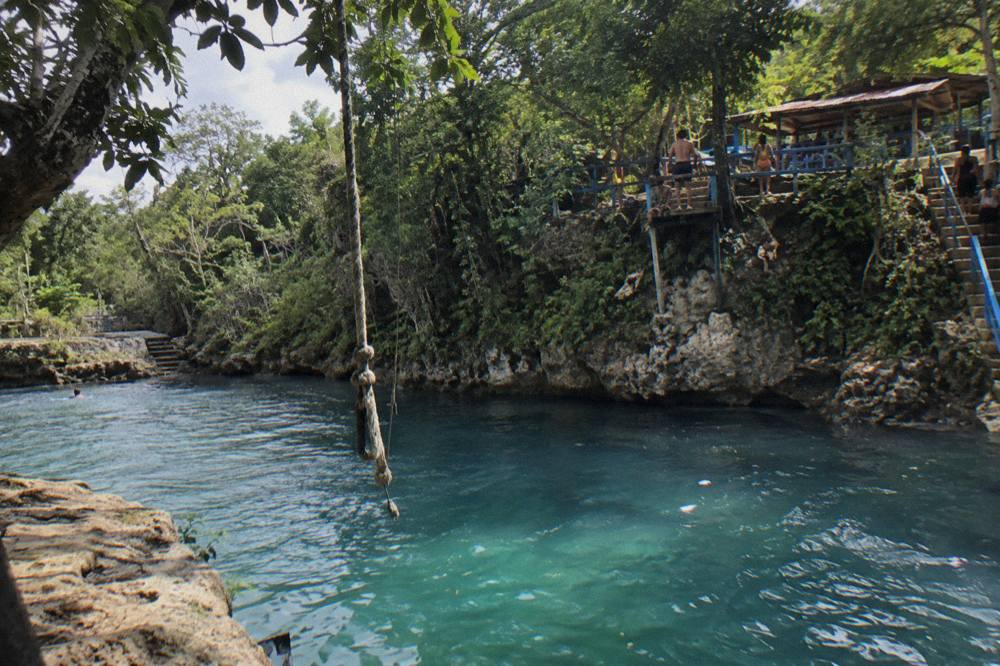 Wooden rope swing hanging over a blue waterfall pool in Jamaican jungle