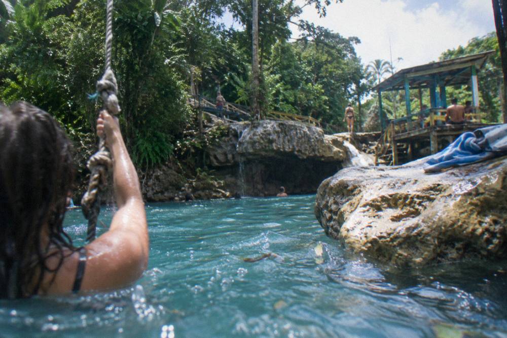 Swimmer holding a rope swing above a clear blue pool at a Jamaican waterfall