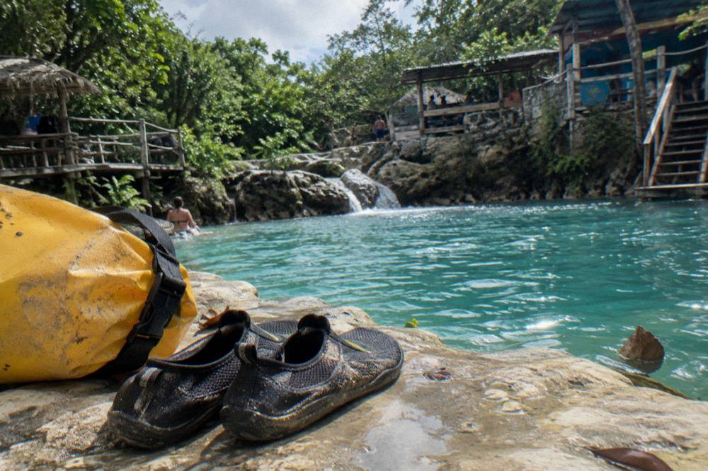 Pair of water shoes and a dry bag resting on a rock beside a turquoise pool