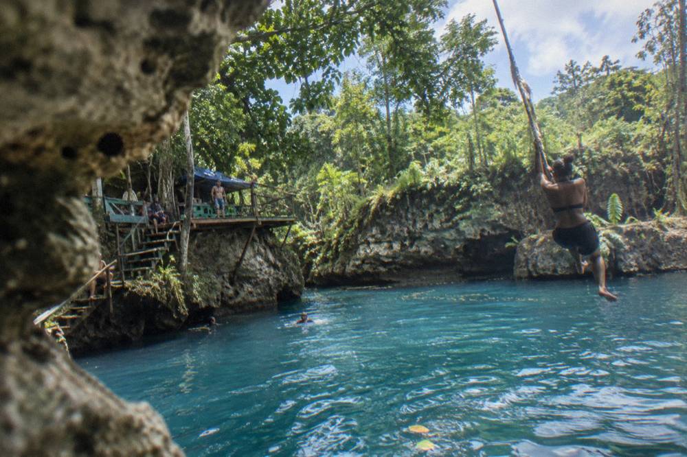 Swimmer on a rope swing above a blue limestone pool surrounded by tropical trees