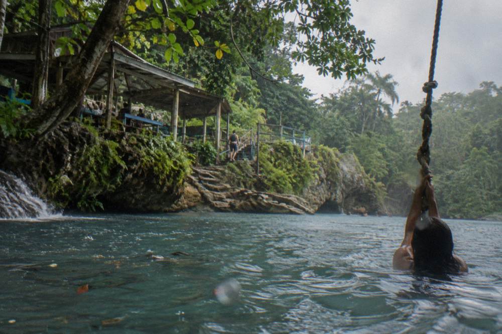 Swimmer holding a rope swing above a clear blue pool beneath jungle canopy in Jamaica