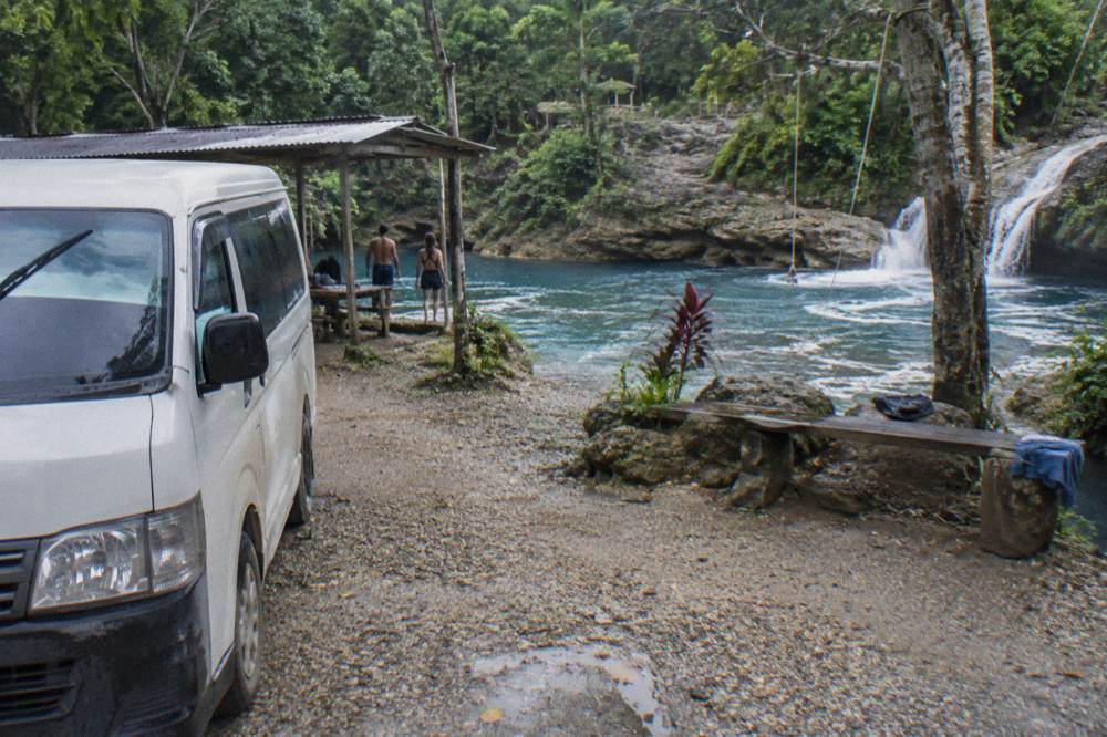 Tour van parked on a gravel road beside tropical forest near a waterfall entrance
