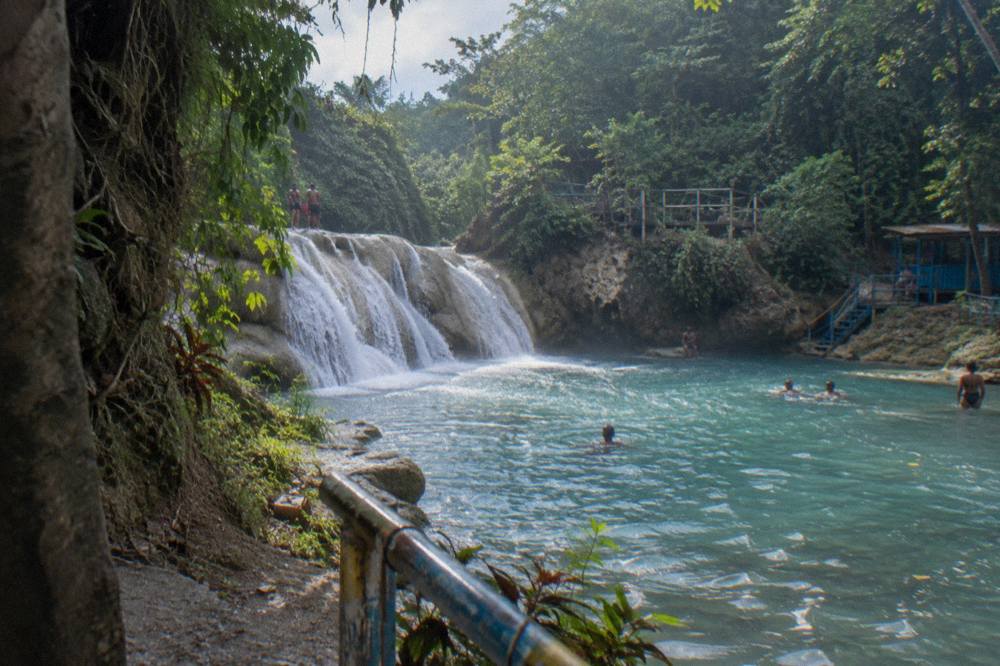 A wide view of cascading waterfalls pouring into a blue limestone pool surrounded by dense green forest
