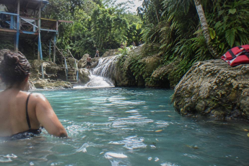 Swimmer entering a blue limestone pool at the base of a jungle waterfall
