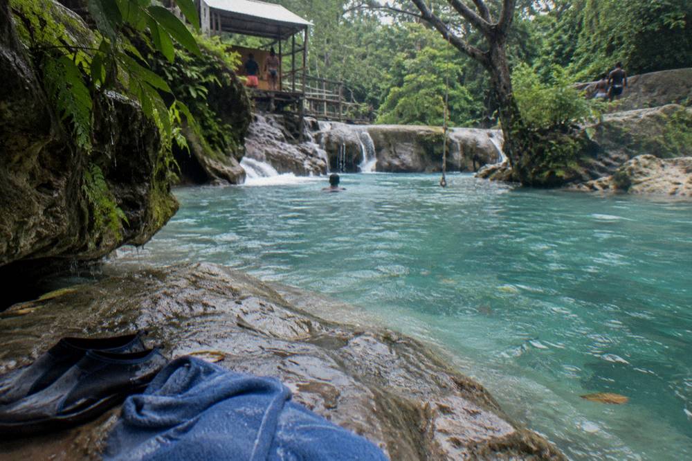 Water shoes and towels set on a rock beside a turquoise swimming hole