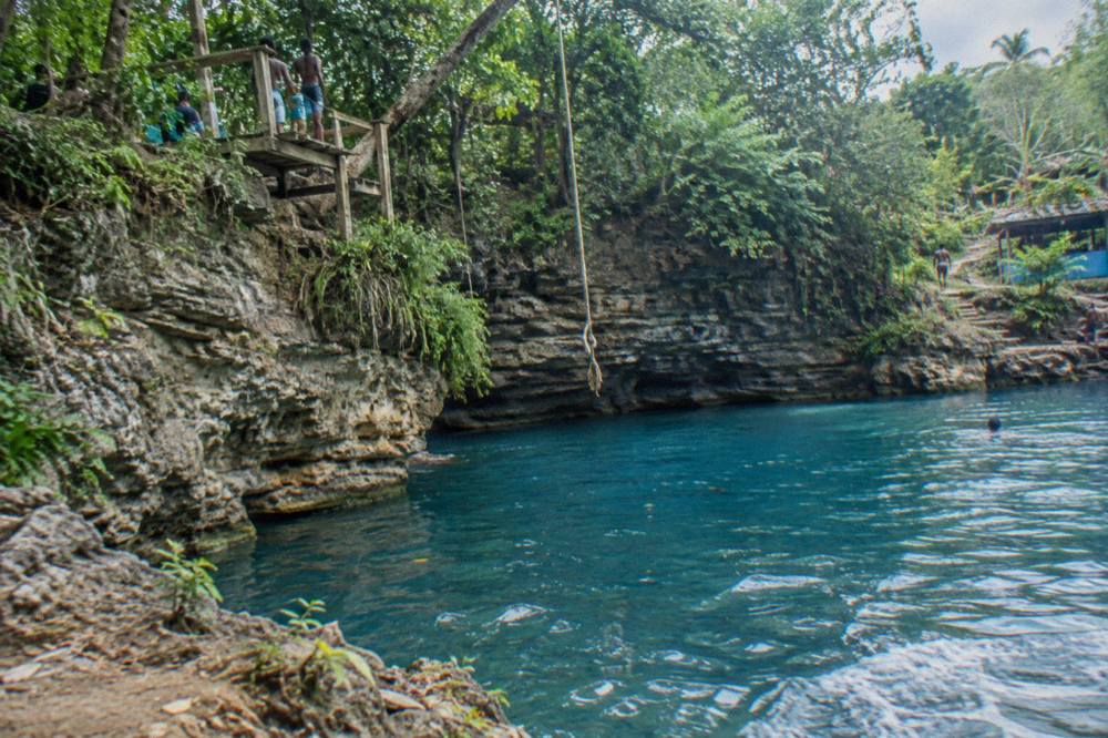 A jungle river gorge with layered limestone rocks and a rope swing hanging over blue water