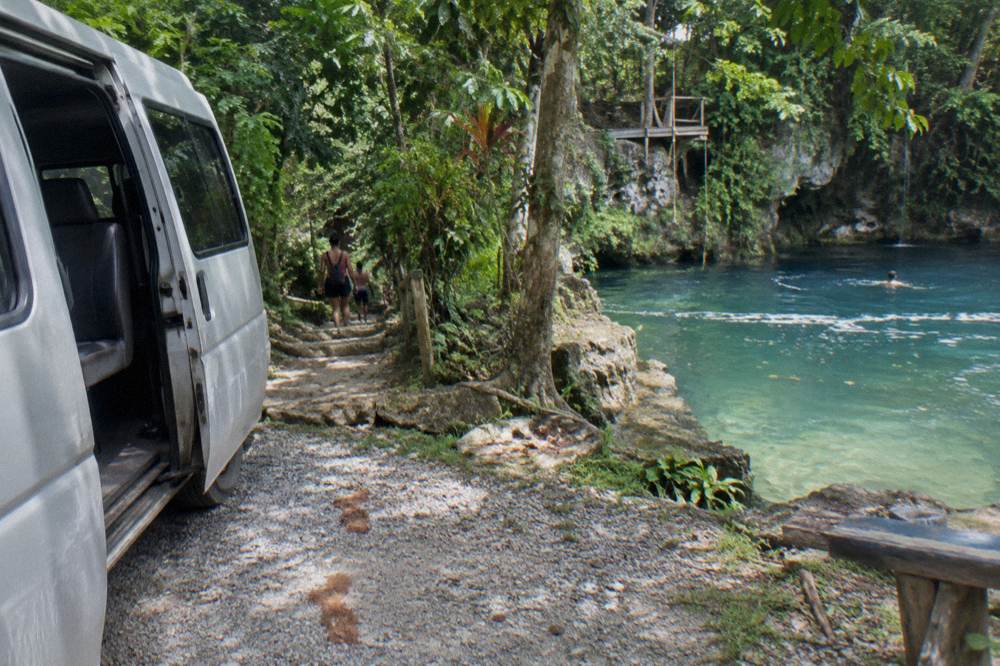 A parked tour van beside a jungle trail entrance at a Jamaican waterfall attraction